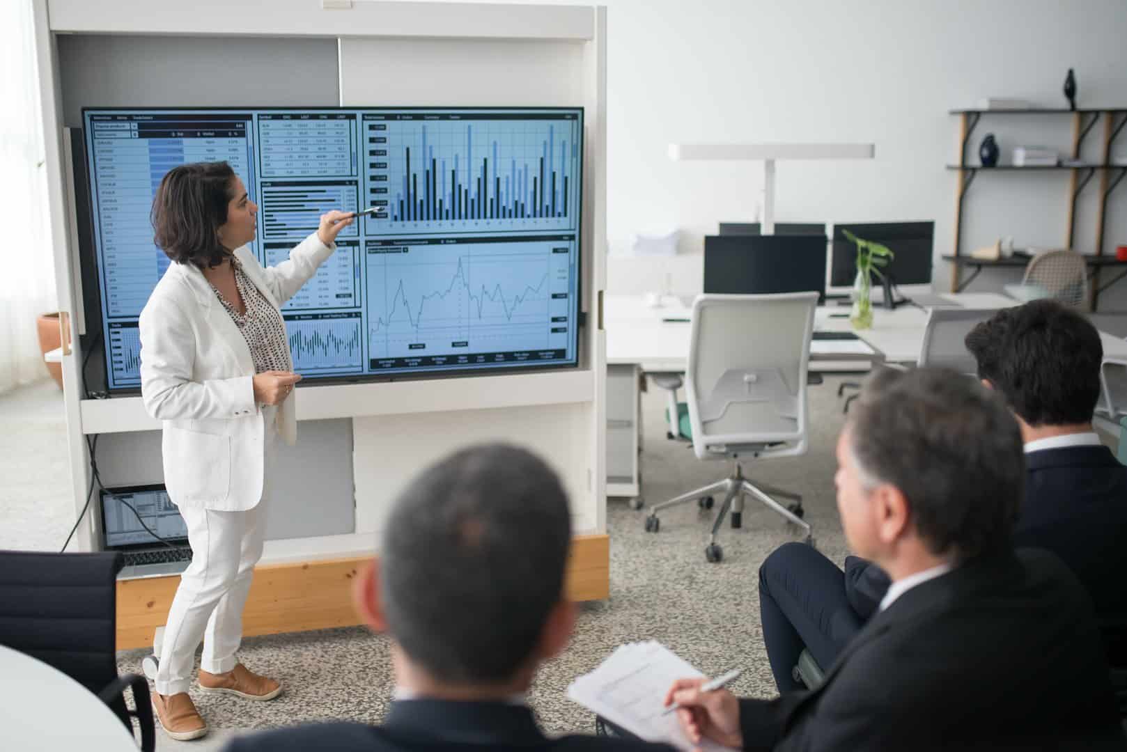 In a board room, a formally dressed woman points at a statistics presentation to the audience in front of her, implying data and certainty.