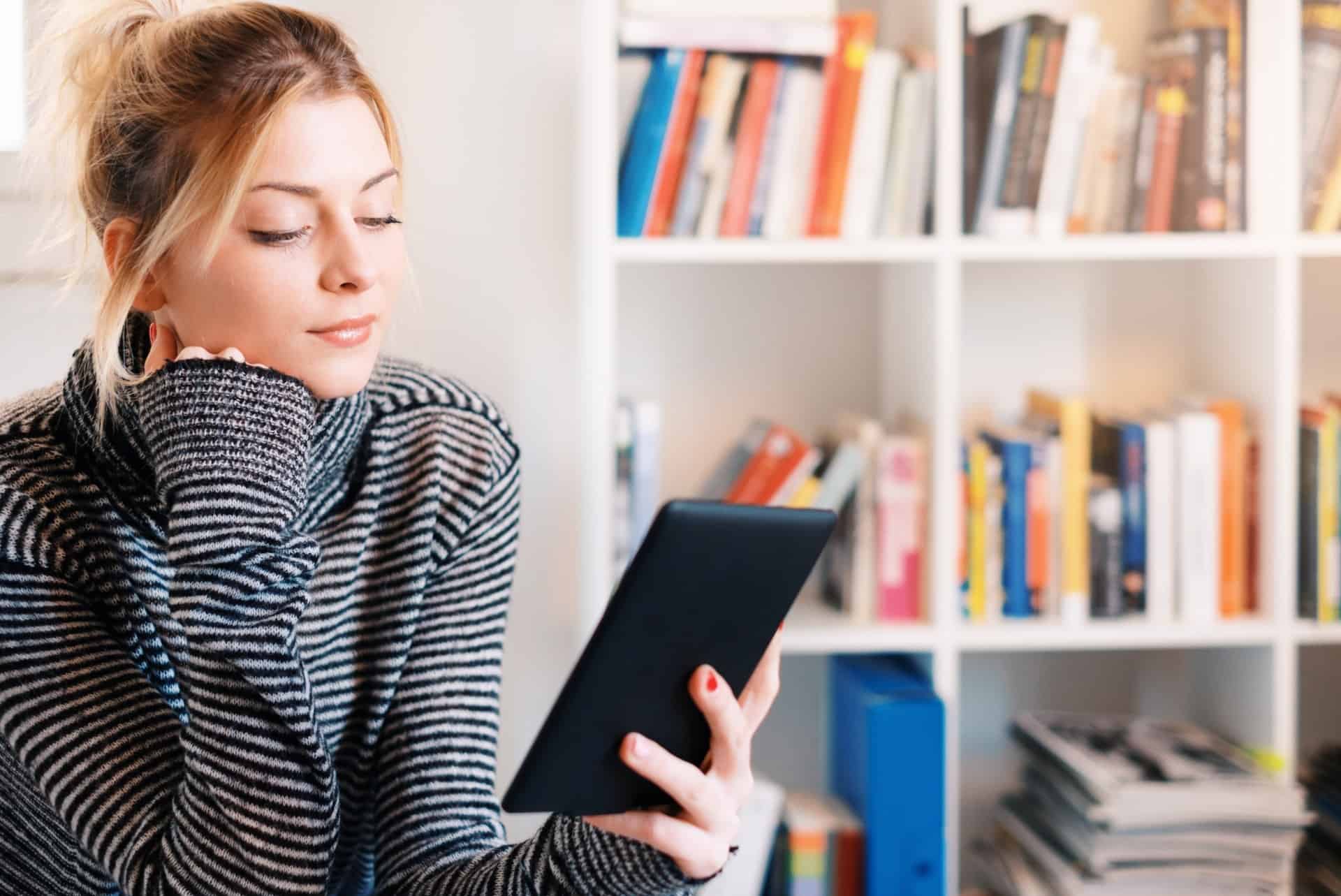 A woman reads from a handheld device with books arranged on a bookshelf behind her, implying book publishing.