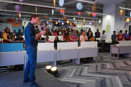 A man stands on the dais and speaks on a microphone in front of other employees during an event at the workplace.