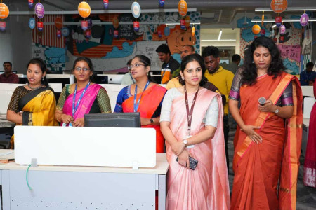 A photograph shows five women employees standing next to each other behind a desk during an event at the workplace. The woman at the extreme right holds a mic and looks at it.