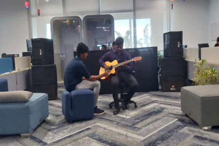 An employee plays guitar as he sits in a chair in the relaxation space in the office. Another man sits beside him and holds a microphone near the guitar.